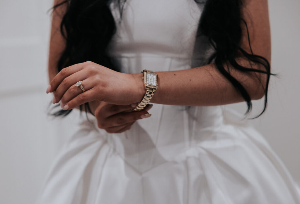 A close-up shot of a bride's hand wearing a diamond engagement ring, with her wrist adorned by a gold watch. She is in a white bridal gown, with her hand slightly raised as she adjusts her watch. The focus is on the jewelry and the bride's delicate, natural pose.