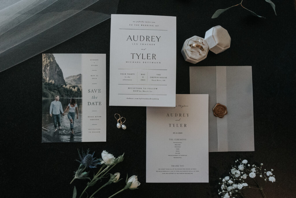 Flat lay of wedding stationery, featuring a "Save the Date" card, a formal wedding invitation with details about the ceremony, and a matching envelope sealed with a wax stamp. The photo also includes a pair of pearl earrings, two wedding rings in a white hexagonal box, and a sprig of flowers on a dark surface.