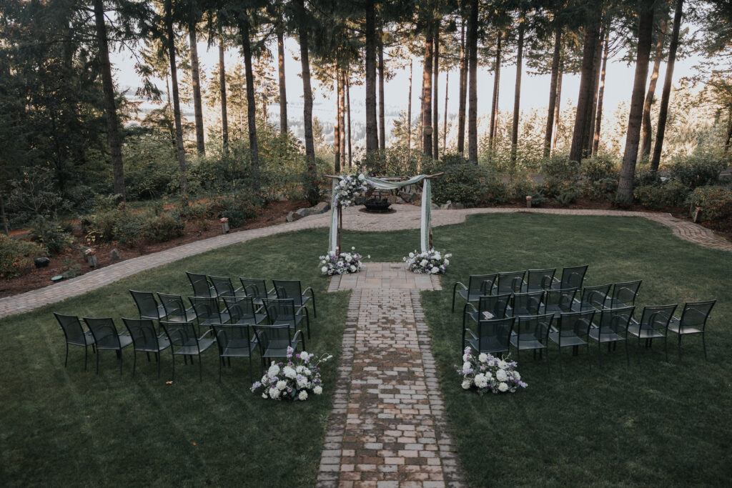 A serene outdoor wedding ceremony setup with a flower-adorned arch, white chairs arranged along a stone path, and lush greenery in the background.