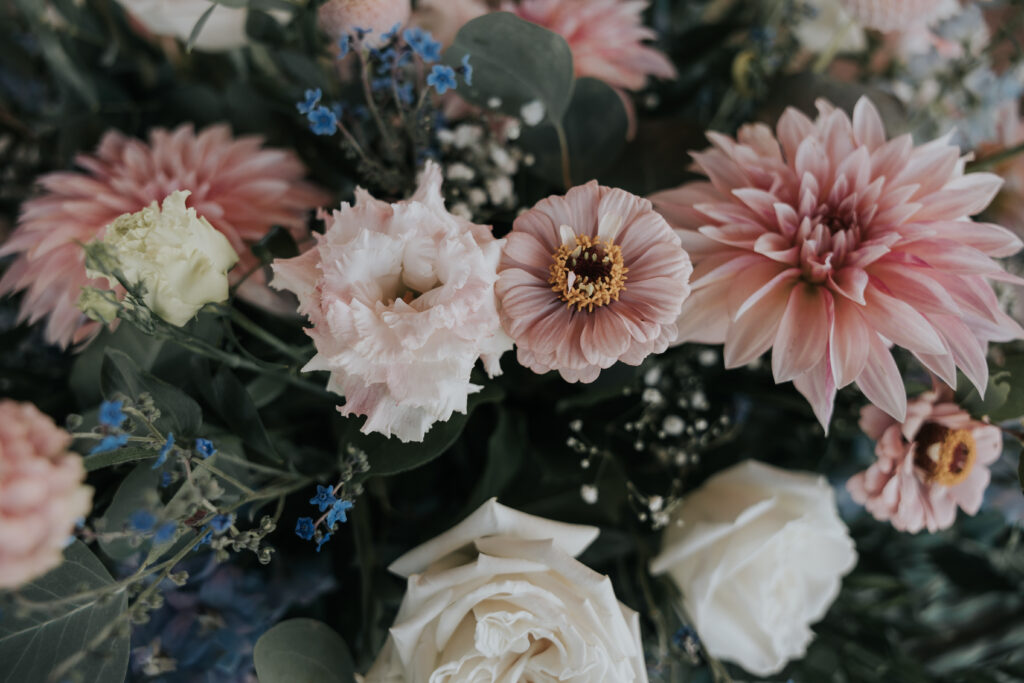 A close-up of a colorful bouquet featuring soft pink dahlias, pale pink lisianthus flowers, white roses, and small blue forget-me-nots, with delicate greenery and small white baby's breath accents.