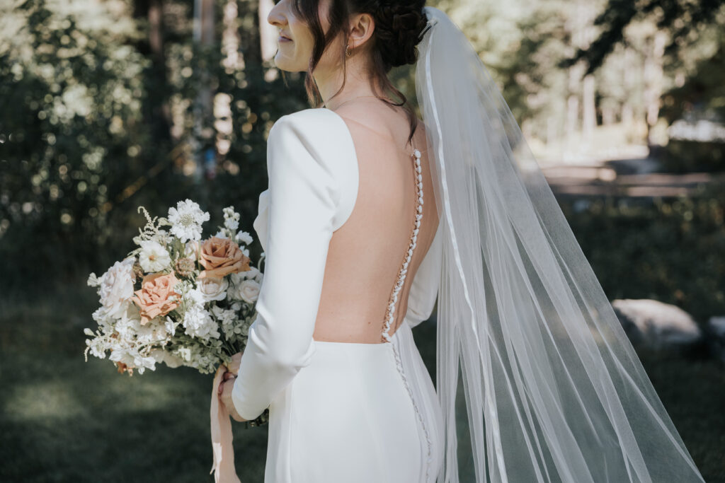 Close-up of a bride holding a bouquet of blush and white roses, with her back to the camera showing a low-cut wedding dress with delicate button detailing. A flowing veil cascades down her back as she smiles in a soft, natural setting.