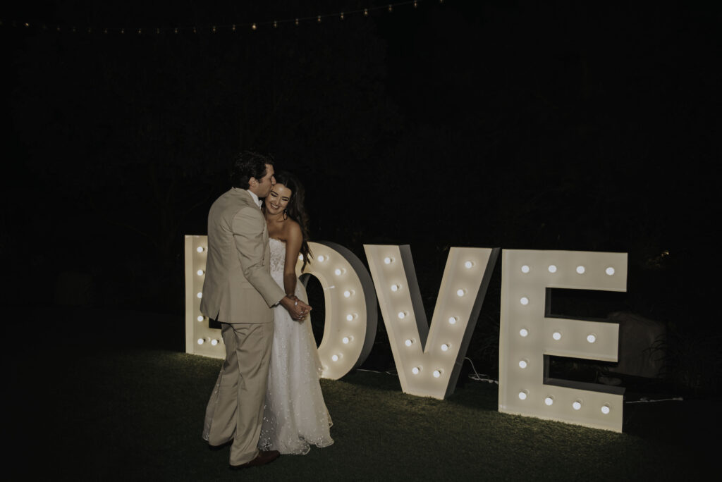 Bride and groom sharing a sweet moment in front of a glowing LOVE marquee sign at night, a creative twist on modern wedding send-off ideas.