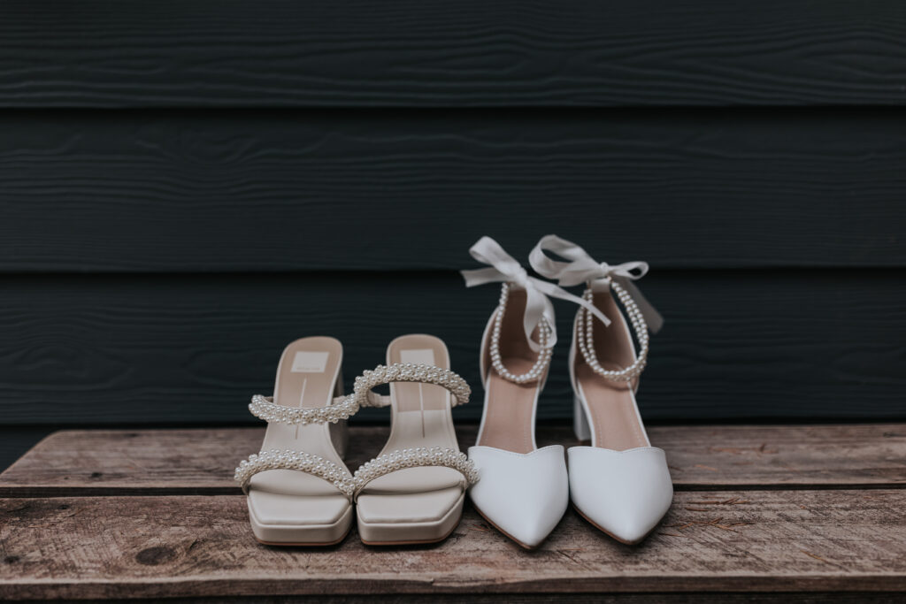 Two pairs of white bridal shoes placed side by side on a wooden surface. On the left, a pair of flat sandals with pearl detailing on the straps. On the right, a pair of pointed-toe heels with pearl embellishments and satin ribbons tied around the ankle. The background is a dark wooden panel.